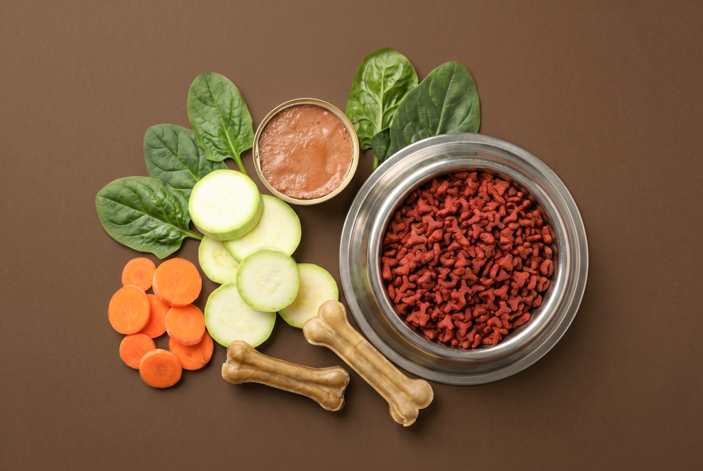 Top-down flat lay of a stainless steel bowl filled with dry dog kibble, alongside an open tin of wet dog food, fresh spinach leaves, sliced courgette, carrot rounds, and two natural chew bones, arranged on a warm brown background with ample copy space.