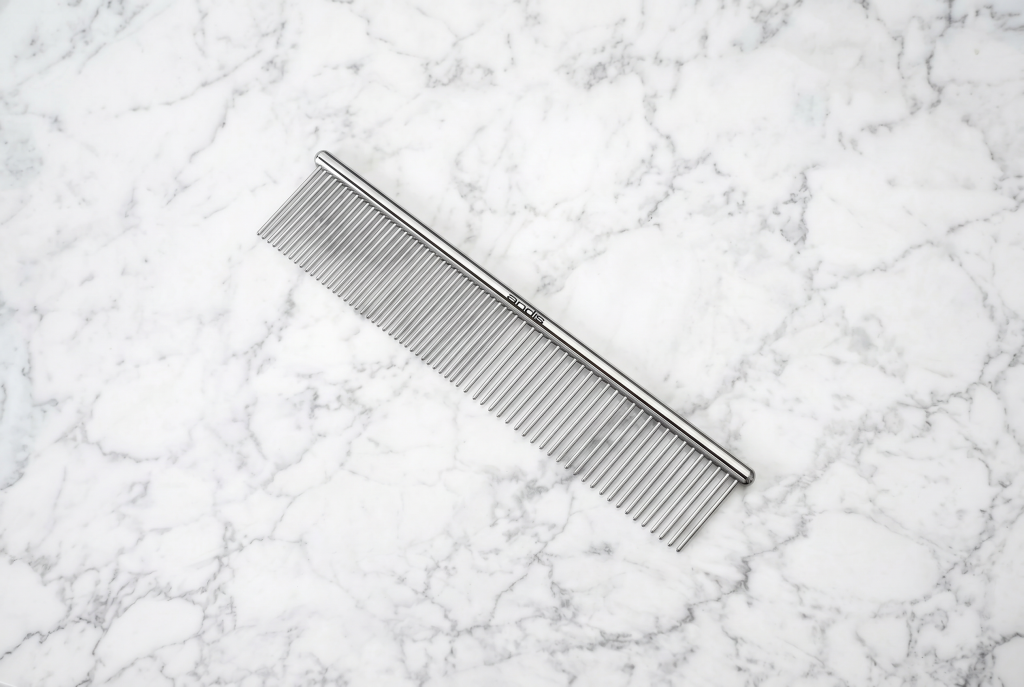 Metal dog grooming comb with rounded stainless steel teeth, featuring both wide- and fine-spaced sections, displayed diagonally on a white marble background.