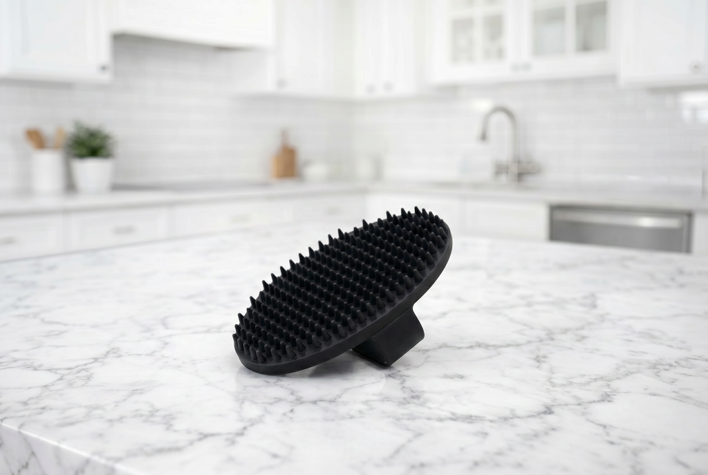 Black rubber dog grooming brush with soft flexible bristles standing on a white marble kitchen countertop, with a bright, blurred modern kitchen in the background.