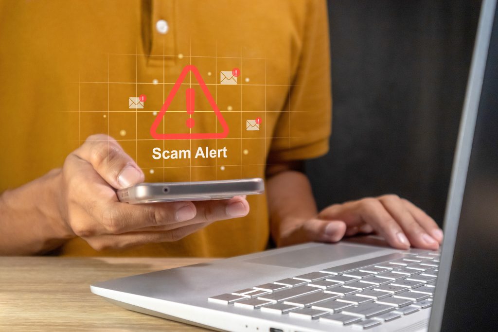 Person holding a smartphone while using a laptop, with a red "Scam Alert" warning symbol and email notification icons overlaid on the screen, representing online scams, phishing, and digital fraud awareness.